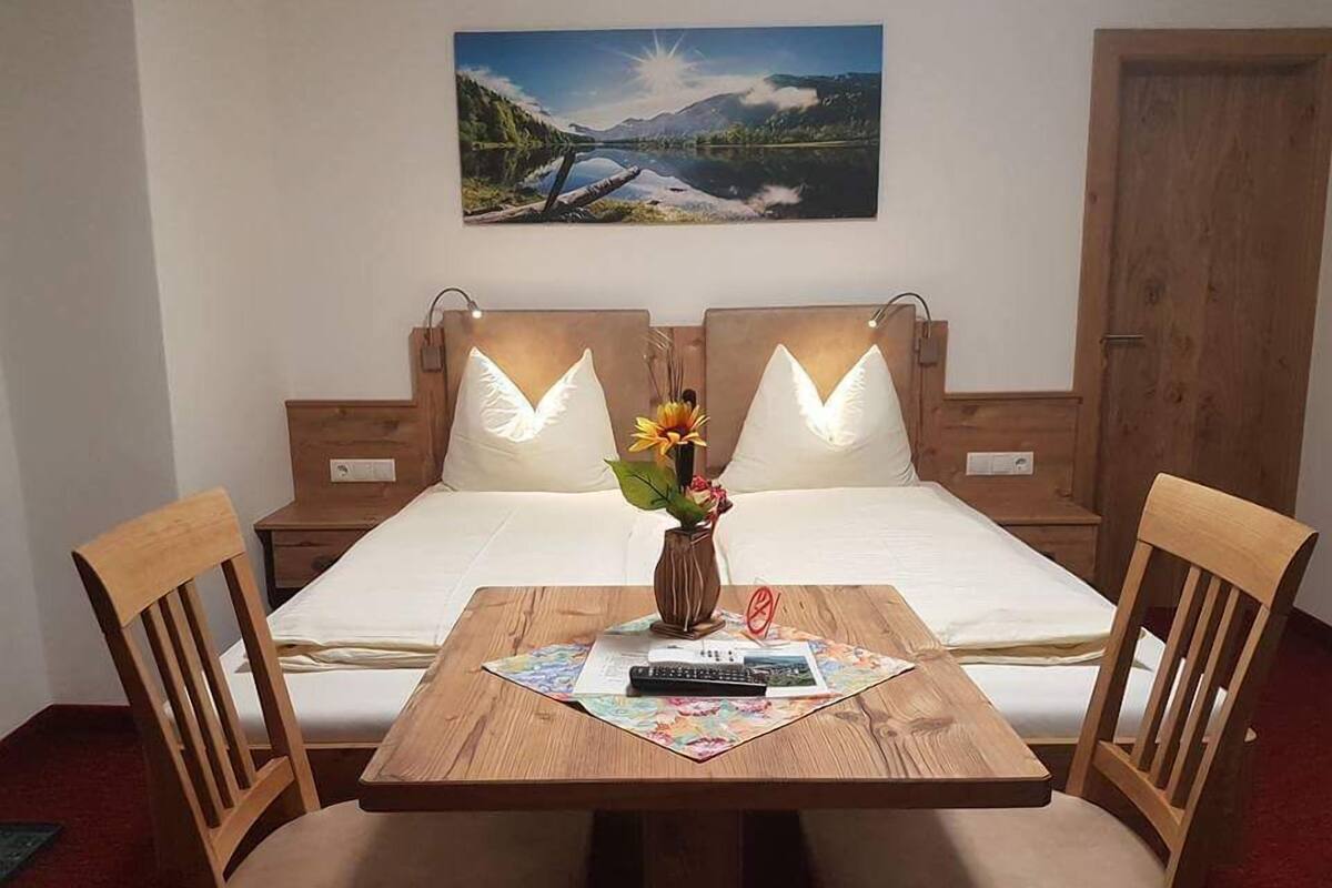 A double bed with a headboard, two pillows, a wooden table with a vase of flowers, and a painting of a landscape on the wall.