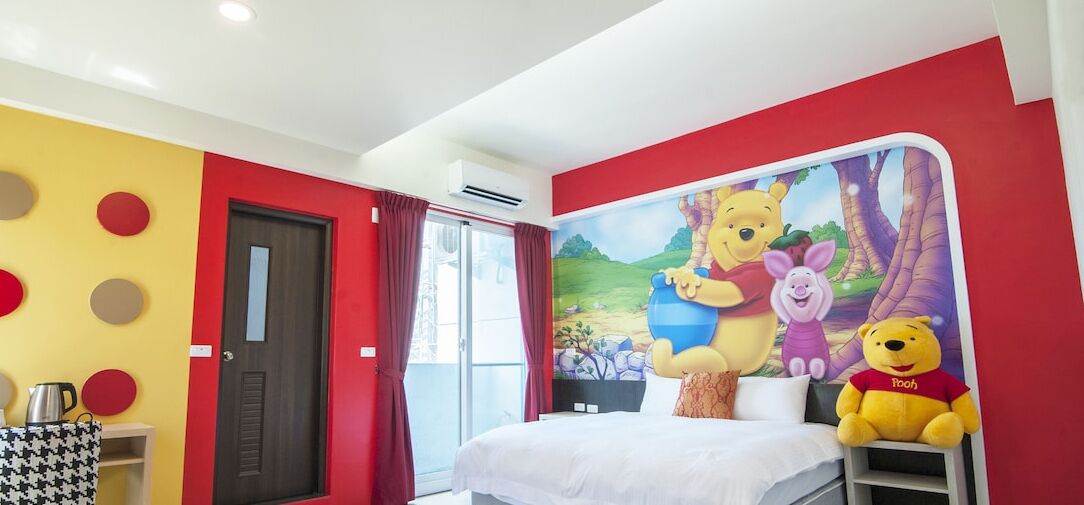 Habitación de hotel con una cama, una ventana con cortinas rojas, una pared con diseños circulares y un peluche de Winnie the Pooh sobre la cama.
