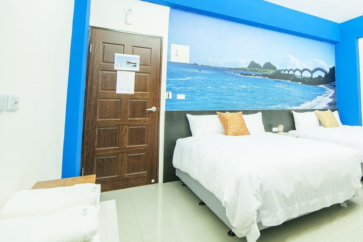 A hotel room with a bed, a door, and a wall mural of a coastal scene.