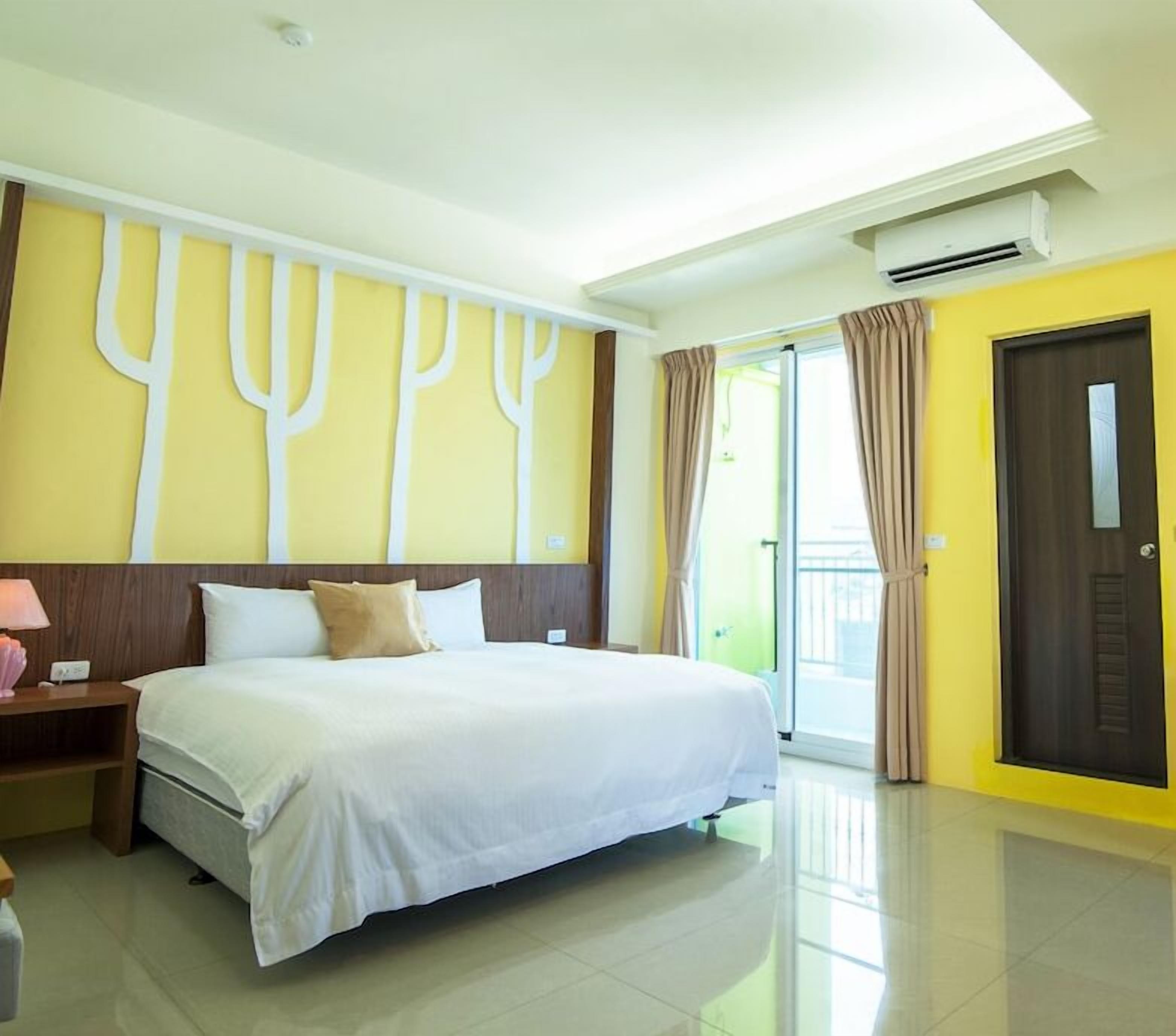 A hotel room with a large bed, a nightstand, a lamp, a window with curtains, and an air conditioning unit.