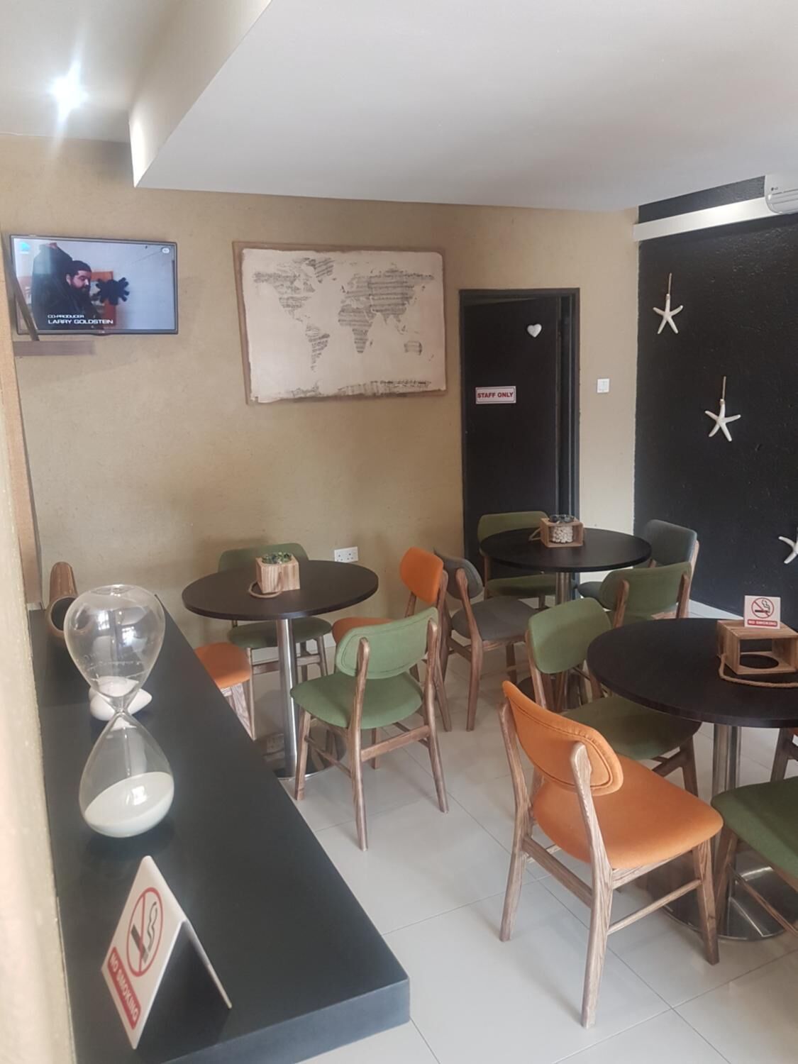 En restaurant med runde borde, grønne og orange stole, et verdenskort på væggen, en tv-skærm og en dør med teksten 'Staff Only'.