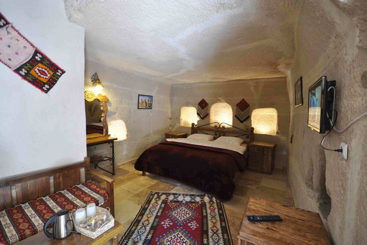 Ein gemütliches Schlafzimmer mit Bett, Nachttischlampen, einer hölzernen Bank, einem kleinen Tisch mit Wasserkocher und Tassen sowie einem gemusterten Teppich.