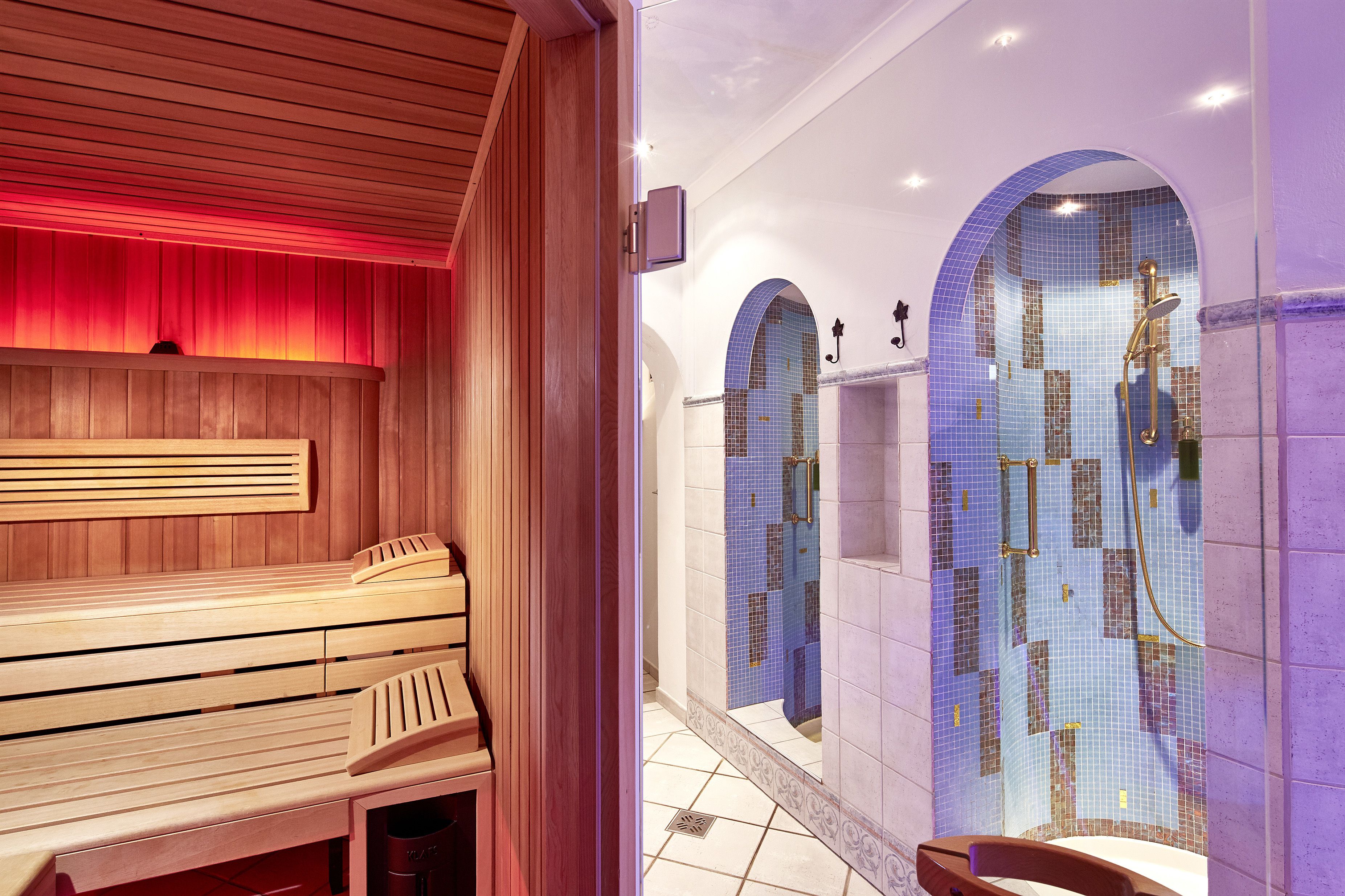 Un espacio de spa con bancos de madera, una pared con azulejos azules de mosaico y una zona de ducha con puerta de vidrio.