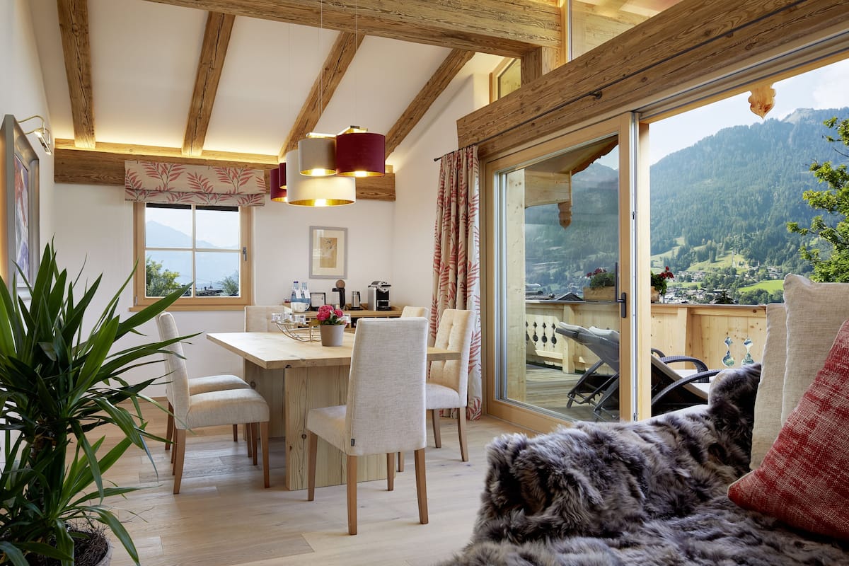 A dining area with a wooden table and chairs, a potted plant, and a view of a mountainous landscape through large windows.