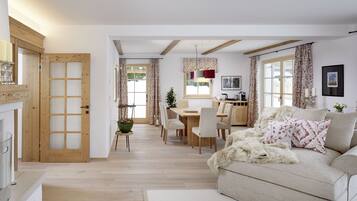 Amplia sala de estar con sofá, mesa de comedor y sillas. Se observa una puerta de madera y una ventana con cortinas.