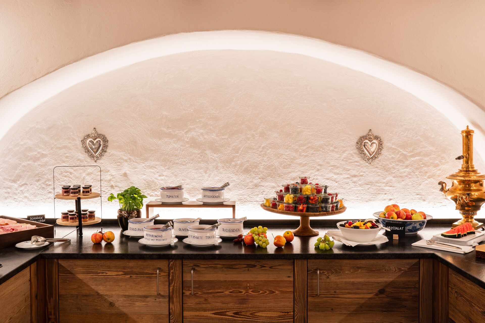 Una encimera de cocina con diversos alimentos y utensilios, incluyendo frascos de condimentos, un frutero y una estufa de bronce decorativa.