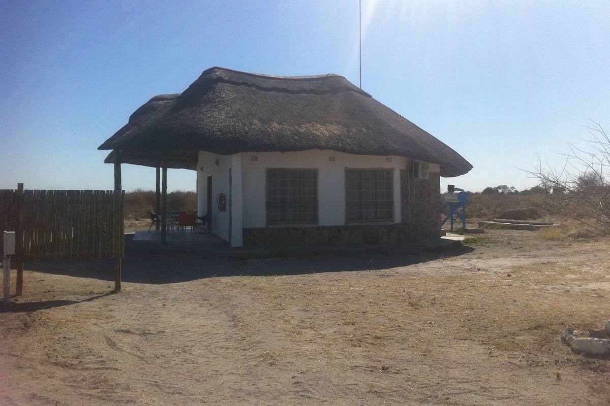A small, single-story building with a thatched roof, a fenced outdoor area, and a dirt ground.