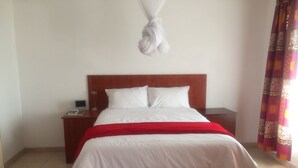 Habitación de hotel con cabecera de madera, cubrecama blanco, manta roja y una mesita de noche con lámpara, además de una alfombra roja.
