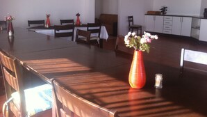 Un salón de conferencias con una larga mesa, varias sillas y jarrones con flores. Hay un cuadro en la pared y una zona de cocina al fondo.
