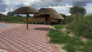 Una construcción con techo de paja y terraza de madera, rodeada de follaje y un camino pavimentado.