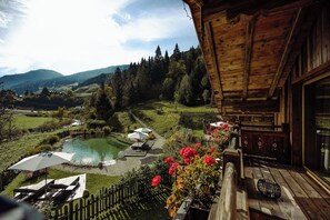 A wooden cabin with a balcony overlooking a pond, surrounded by greenery and mountains.