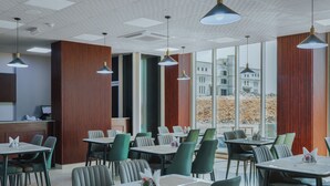 A modern restaurant with multiple dining tables and chairs, large windows offering a view of buildings, and pendant lighting. - Muscat Hills Hotel (Muscat)
