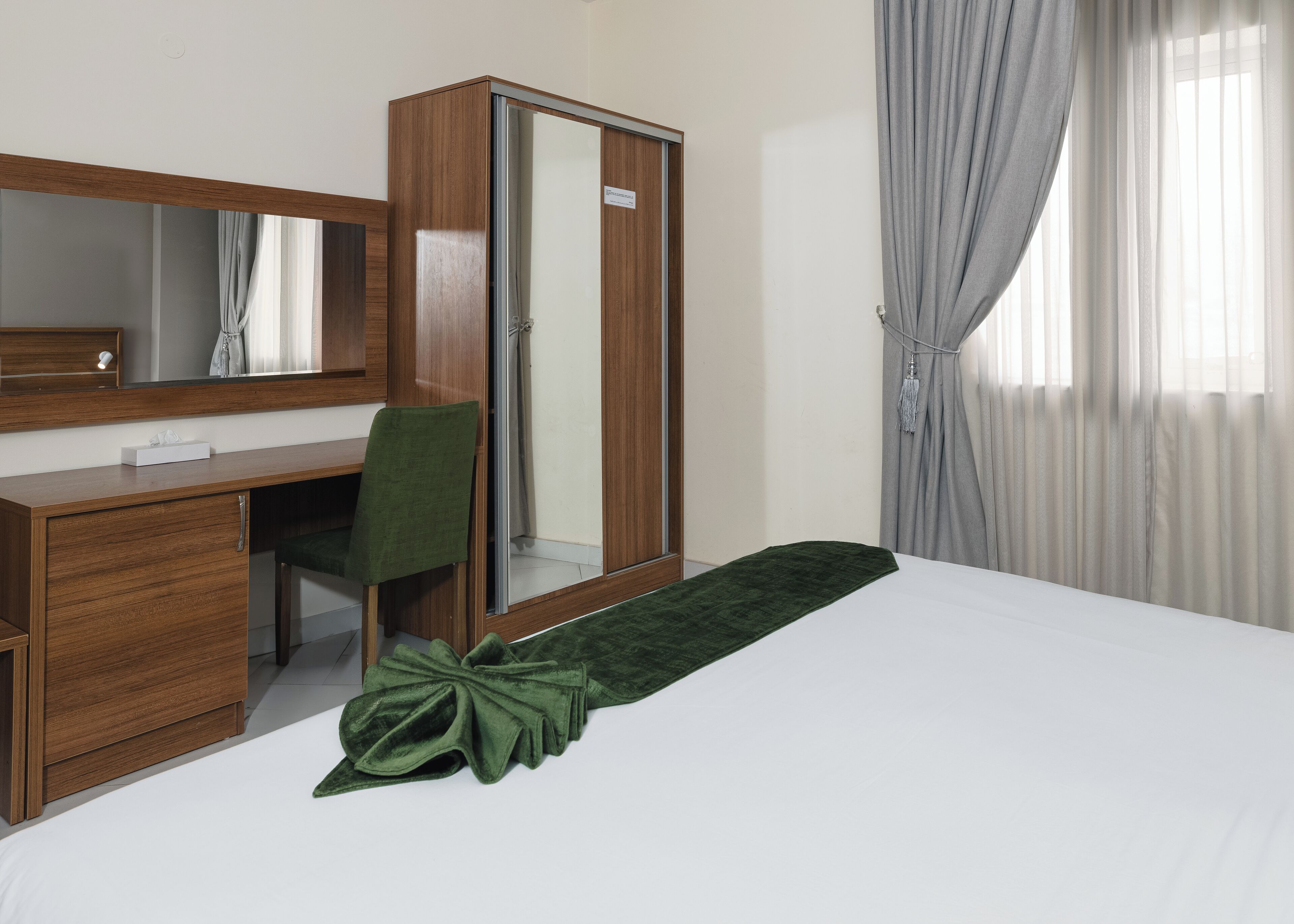 A hotel room with a wooden desk, a green chair, a large mirror, a wardrobe, and a bed with a green throw.