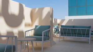 An outdoor seating area with white metal furniture and teal cushions, set against a light-colored wall with a large window. - Muscat Hills Hotel (Muscat)