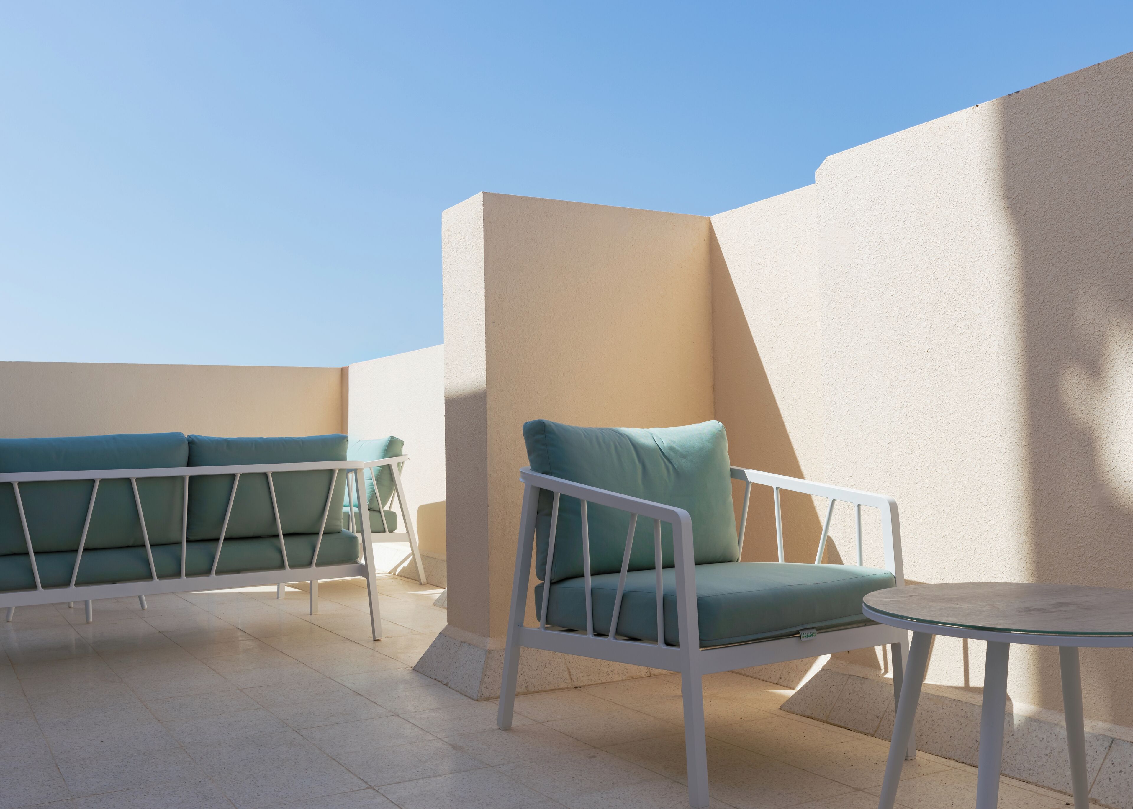 Une terrasse avec deux chaises en métal blanc dotées de coussins verts, une petite table ronde et un mur de couleur beige.