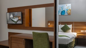 A modern hotel room with a wooden desk, a green chair, a bed with white bedding and green pillows, a large mirror, and a painting on the wall. - Muscat Hills Hotel (Muscat)