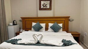 A hotel room with a wooden headboard, two pillows, a bed with white linens, a bedside table with a phone and a kettle, and a small refrigerator. - Al Murooj Hotel Apartments (Muscat)