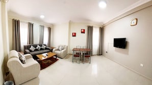A living room with a sofa set, a coffee table, a dining table with chairs, a TV mounted on the wall, and a clock. - Al Murooj Hotel Apartments (Muscat)