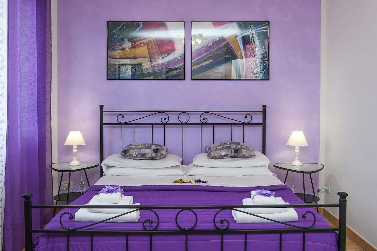 A room with a purple bed, two bedside tables with lamps, and abstract artwork on the wall.