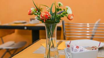 Un florero de vidrio transparente con flores sobre una mesa, un cuenco blanco con objetos, y una silla en una habitación con paredes anaranjadas.