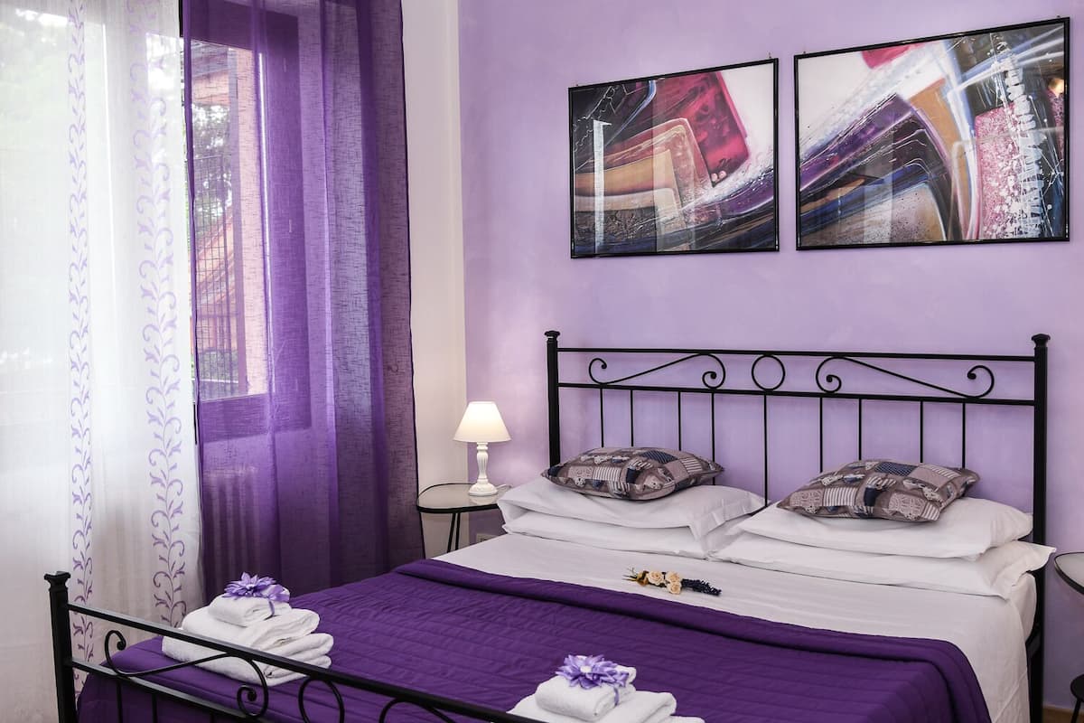 A bedroom with a purple theme, featuring a bed with white and purple bedding, a black metal headboard, and two wall-mounted abstract paintings.