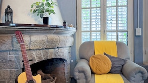 Una sala de estar acogedora con una chimenea de piedra, una guitarra, un sillón cómodo con una manta amarilla y un ventanal con persianas.