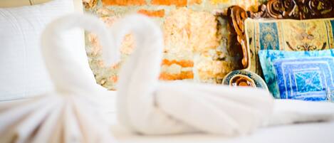 A bed with white linens, a white towel, and a decorative pillow against a brick wall.