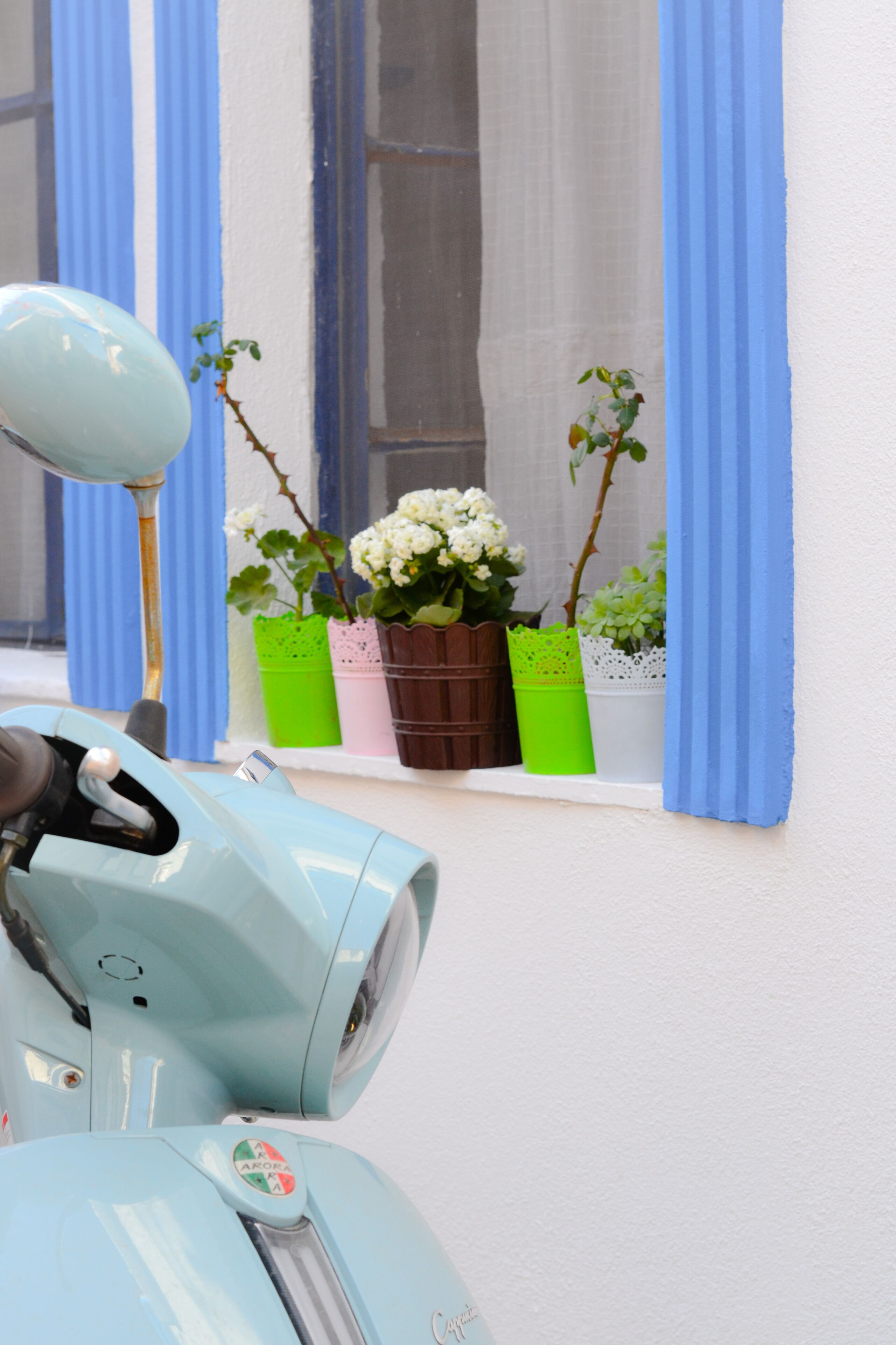 A light blue scooter parked in front of a window with blue curtains.