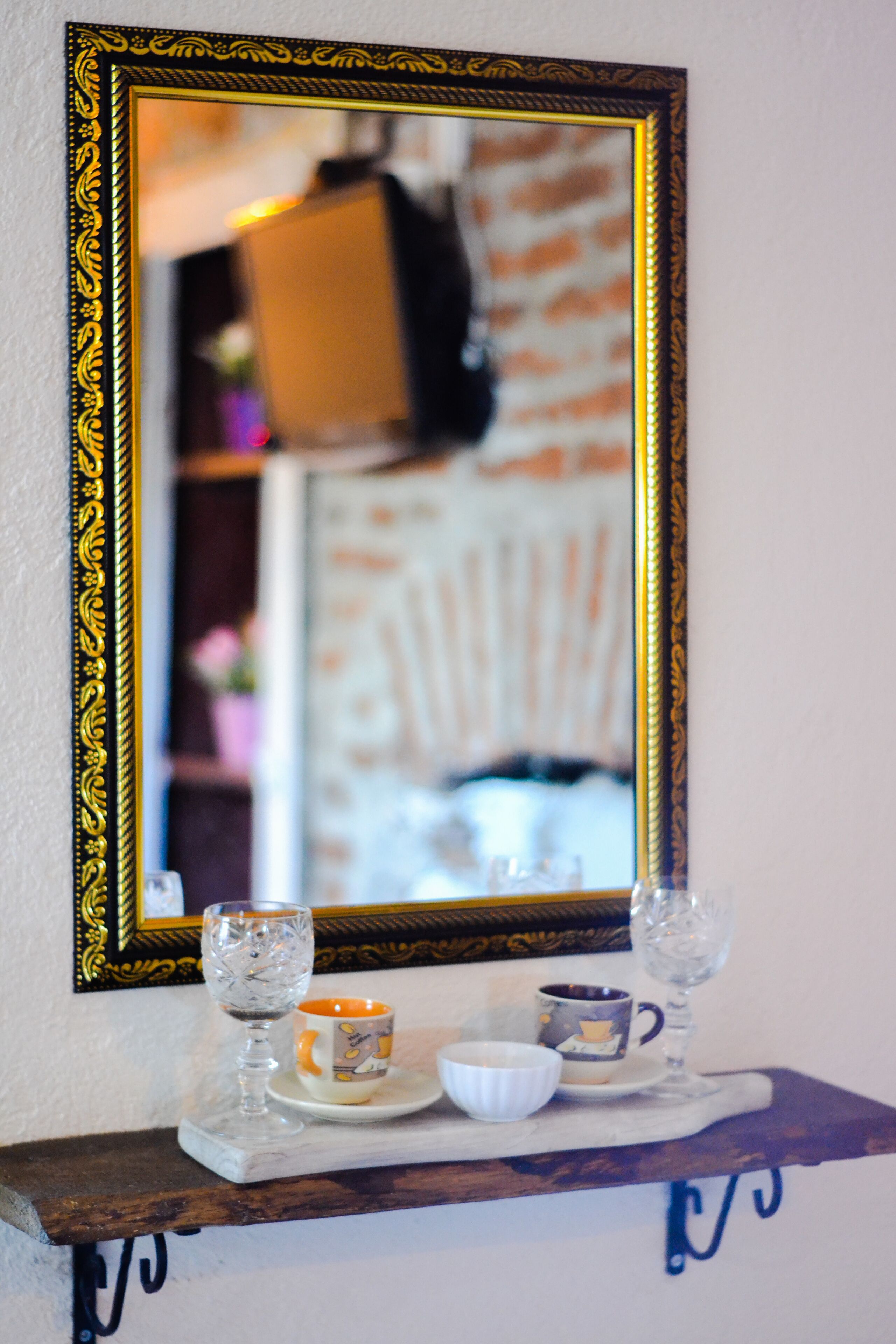 Un espejo con un marco dorado ornamentado, que refleja una habitación con una pared de ladrillo y una estantería con un vaso, una taza y un bol.