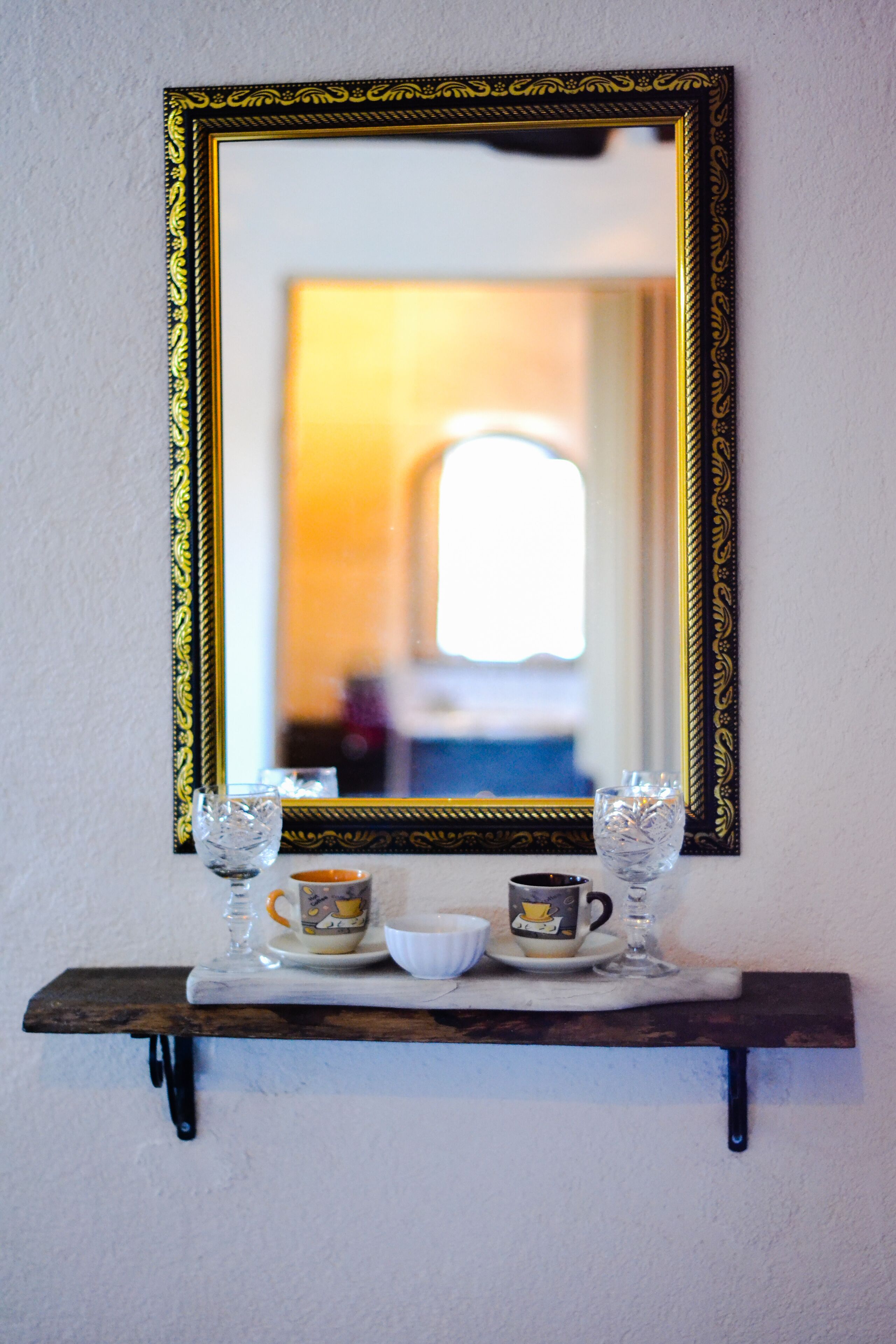 Uno specchio con una cornice dorata decorata su una parete, che riflette una stanza con una finestra e una porta. Sotto lo specchio, uno scaffale in legno contiene una ciotola bianca, una tazza e due bicchieri trasparenti.