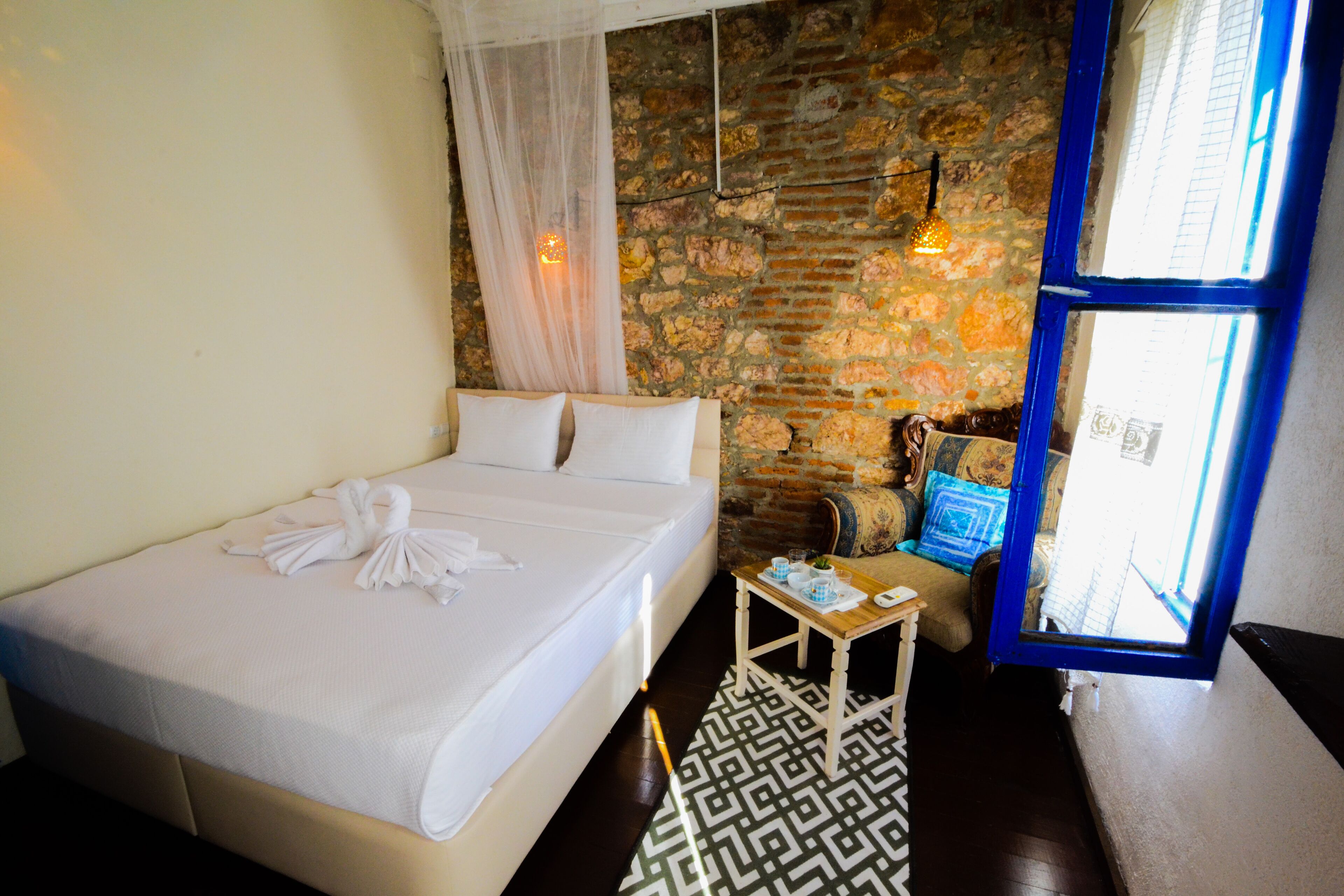A hotel room with a bed, a small table with a book, a window with blue trim, and a stone wall.