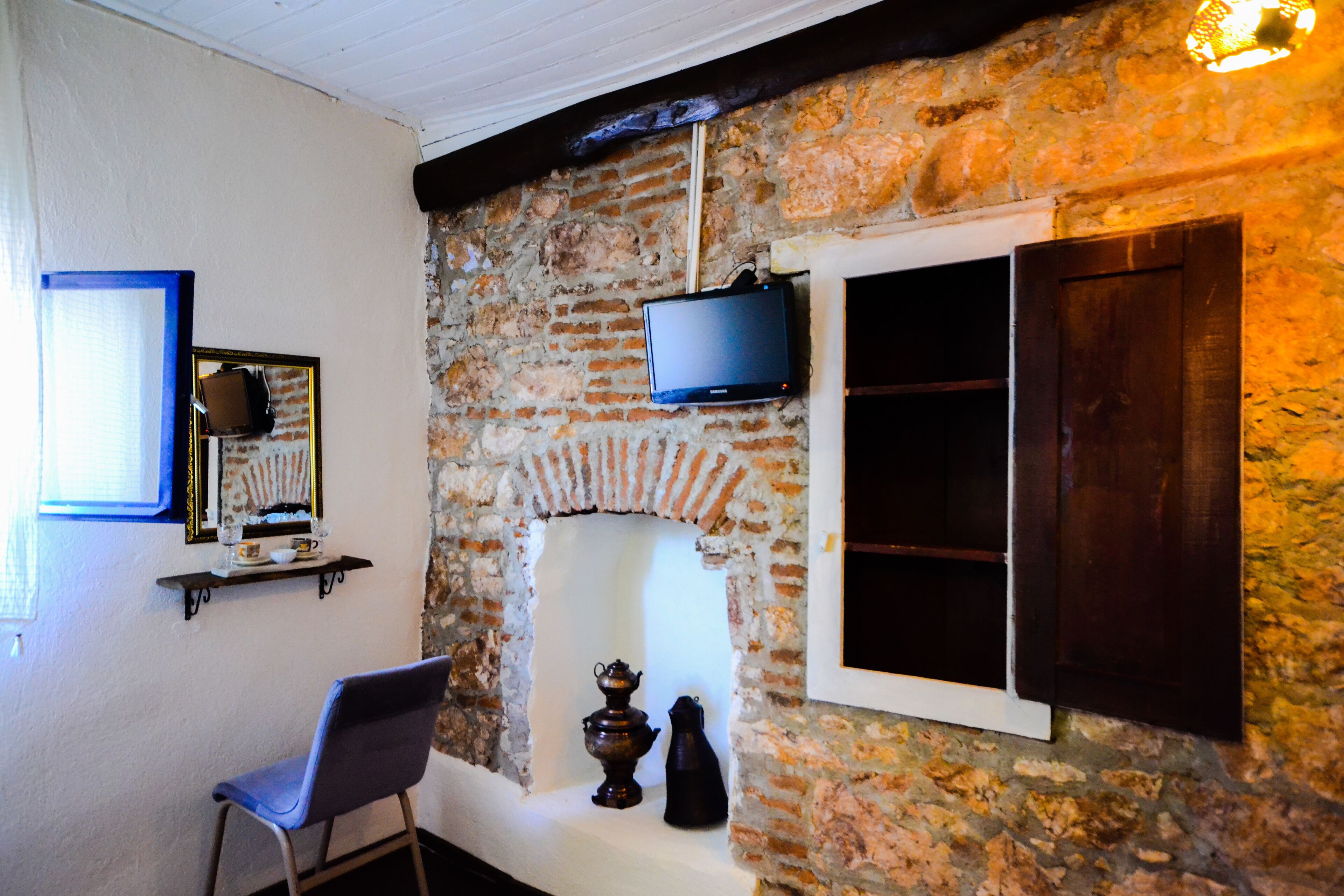 A room with a stone wall, a television mounted above a fireplace, a chair, and a shelf with a mirror and decorative items.