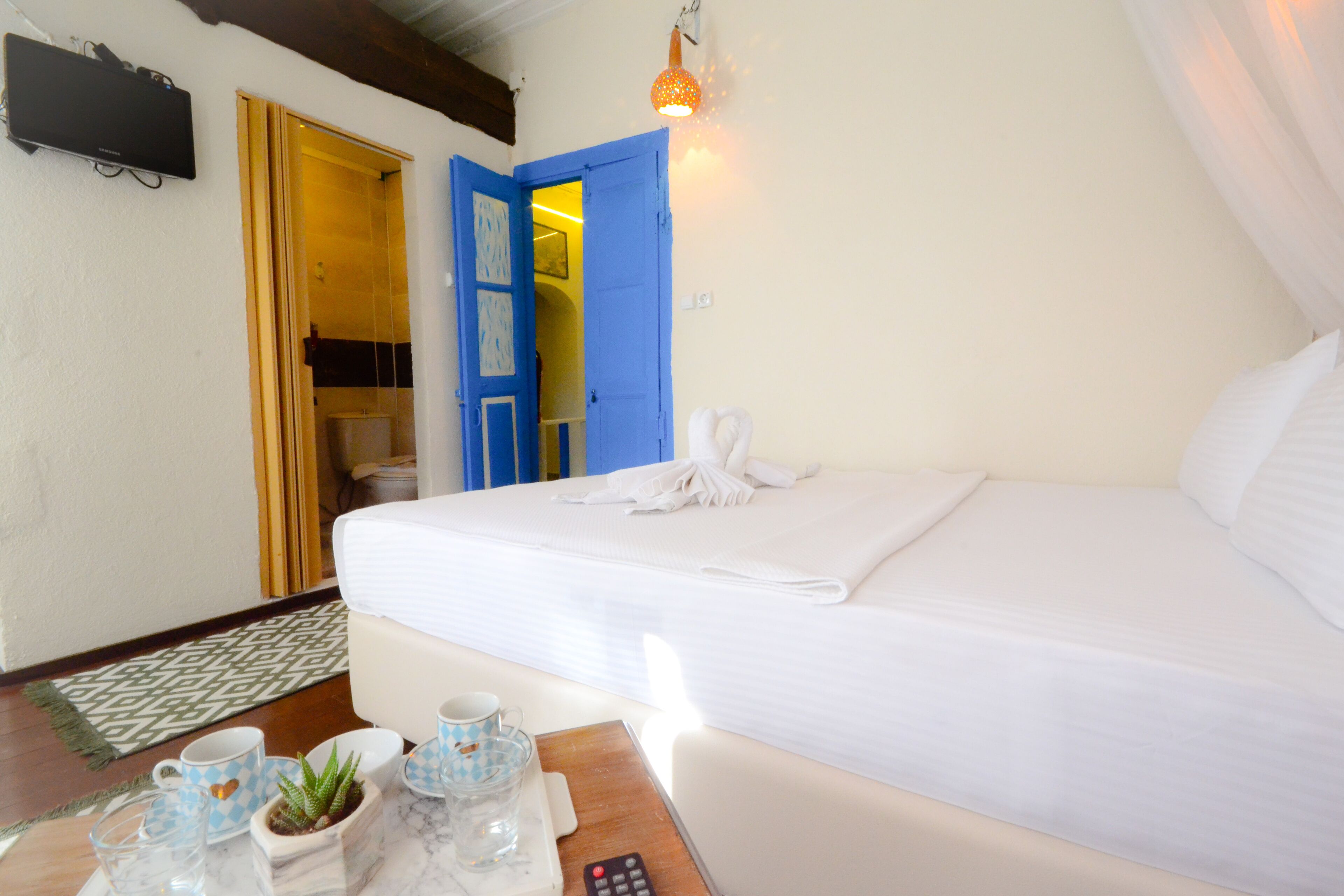 Camera d'albergo con un letto, un tavolo con il telecomando, una pianta e un piccolo tavolo con tazze e piattini.
