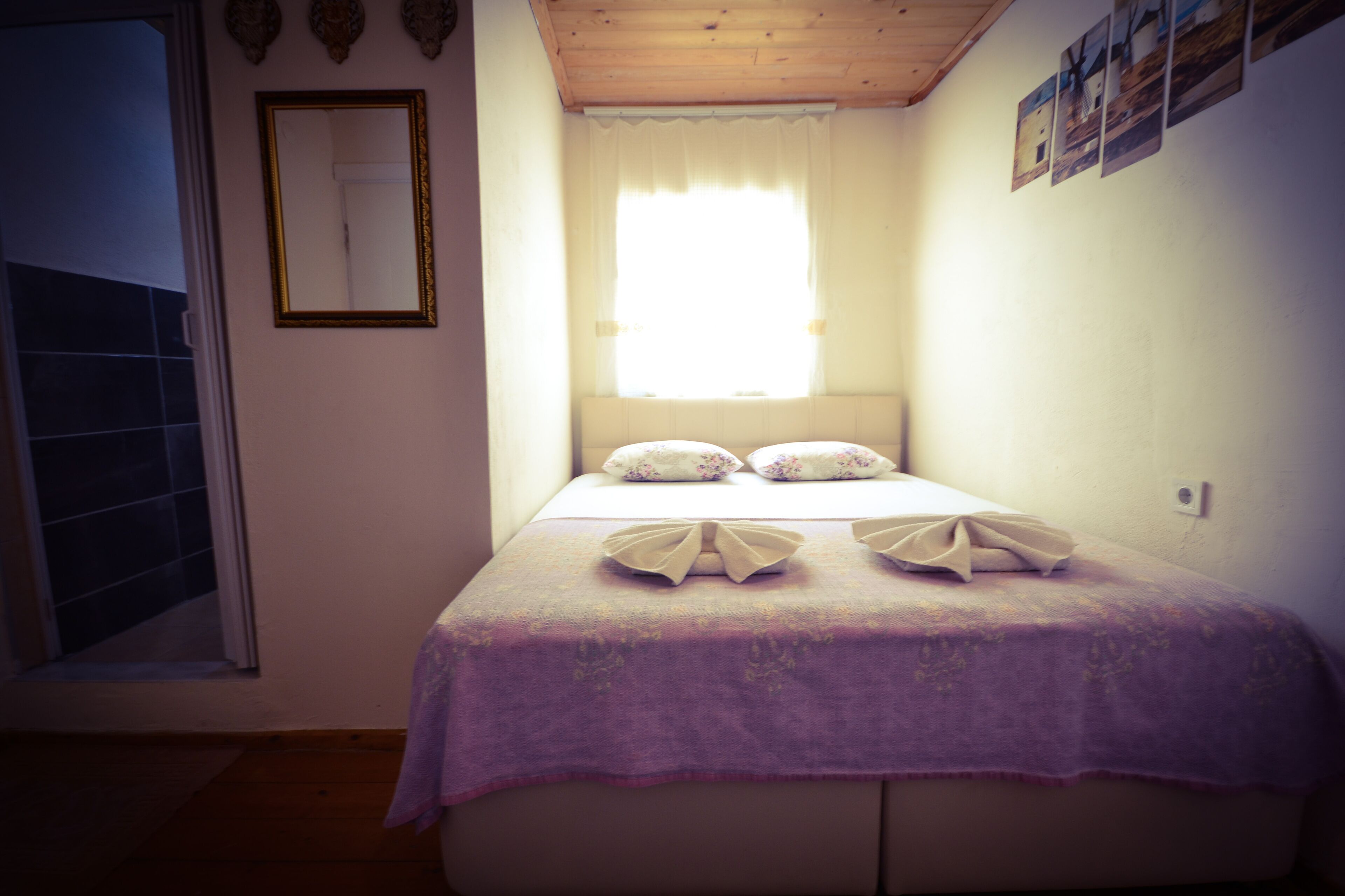 A bedroom with a bed, a window with curtains, a mirror, and a wall with pictures.