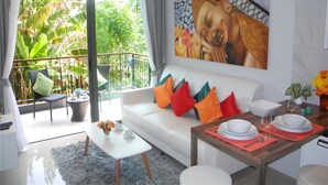 A modern living room with a white sofa, colorful cushions, a dining table set with plates and glasses, and a large Buddha painting on the wall.