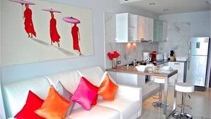 A modern living room with a white sofa, colorful cushions, a coffee table, and a kitchen area with white cabinets and a refrigerator. - Emerald Patong 1 bedroom Modern Apartment (Patong)