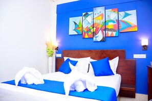 A hotel room with a large bed, blue and white bedding, a wooden headboard, and a wall with colorful abstract art. - Mama Beach Villa (Negombo)