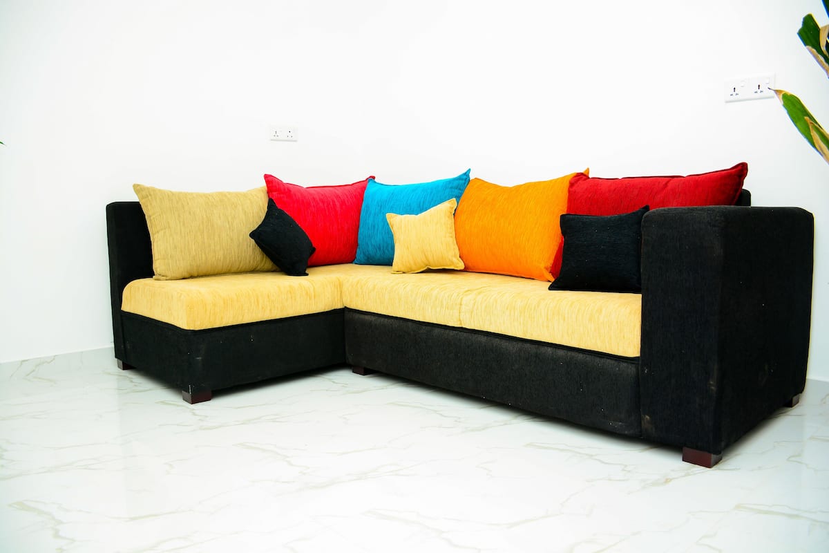 A sectional sofa with a yellow cushion and multiple colorful pillows in a room with white walls and a marble floor.