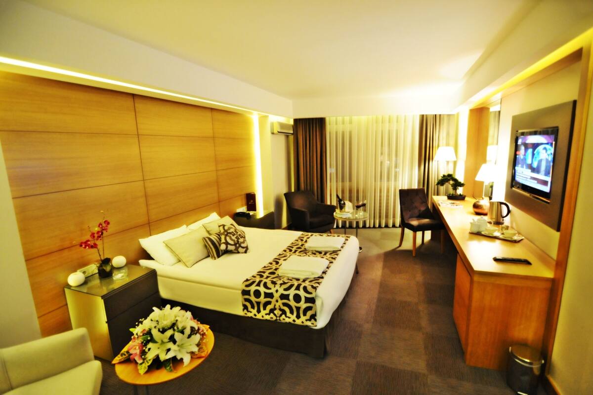 A hotel room with a bed, a desk, a chair, a small table with a lamp, and a television.