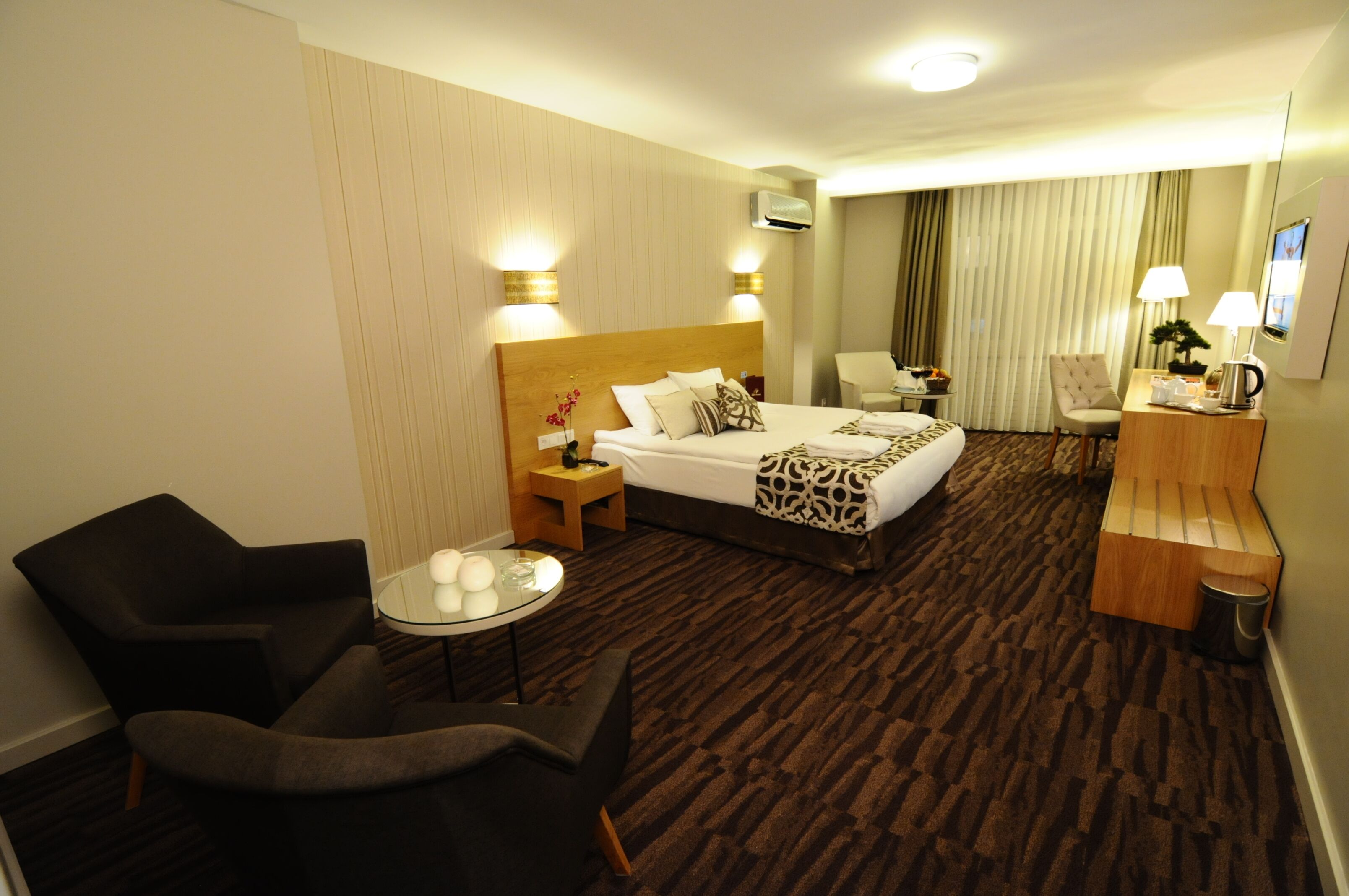 Ein Hotelzimmer mit einem großen Bett, einer Sitzecke mit Stuhl und kleinem Tisch, einem Schreibtisch mit Lampe und einem großen Fenster mit Vorhängen.