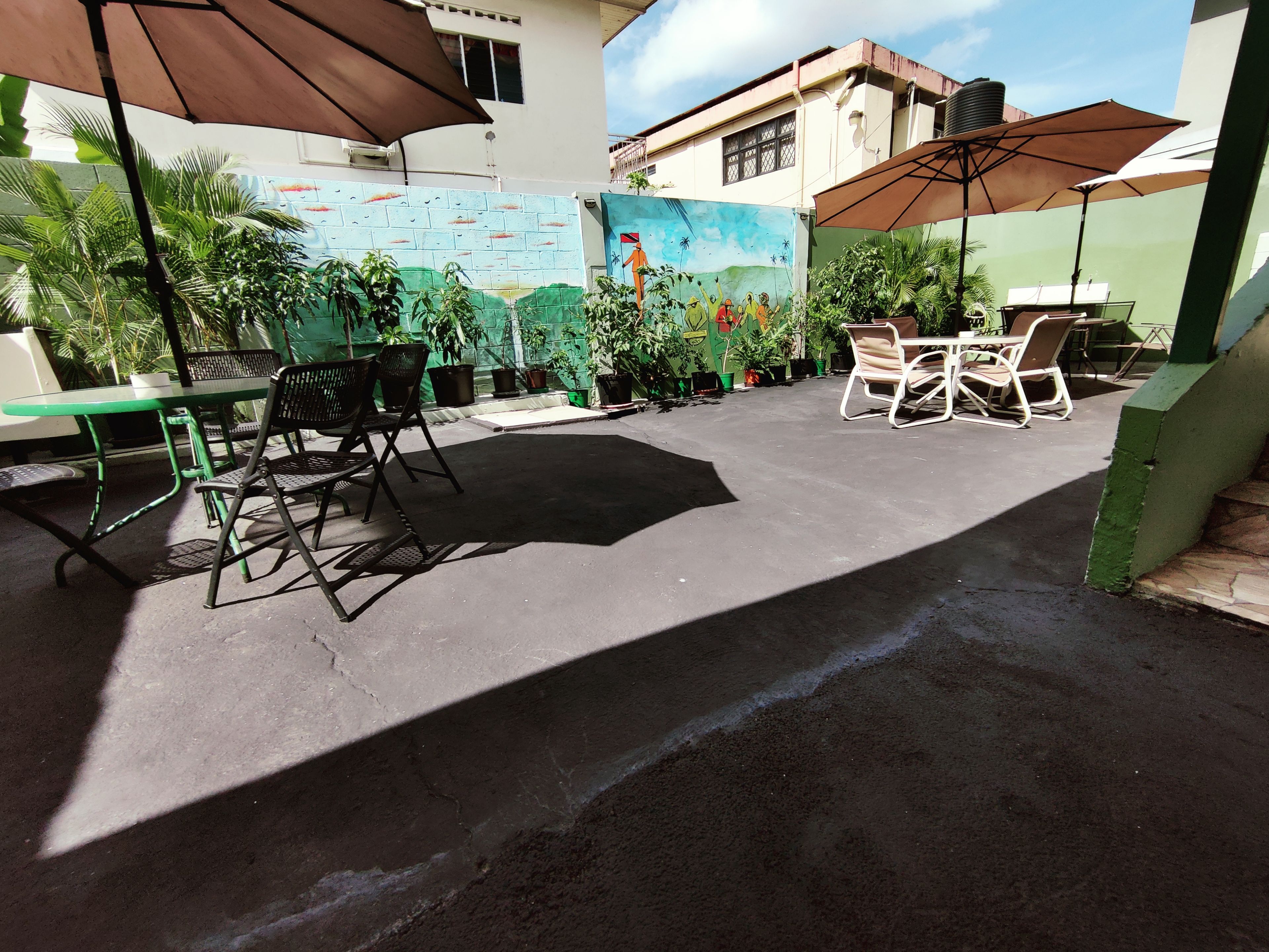 An outdoor patio area with tables and chairs, surrounded by potted plants and a mural on the wall.