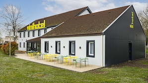 A modern building with a 'vinihotel' sign, featuring a large patio with yellow chairs and tables, and a dark grey extension with a door. - Vinihôtel (Beaune)