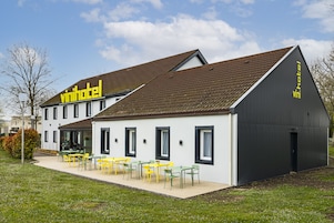Un edificio moderno con un letrero que dice 'vinihotel', que presenta un patio amplio con sillas y mesas amarillas, y una extensión de color gris oscuro con una puerta.