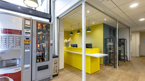 A modern indoor setting with a vending machine, a bright yellow counter, and a display of beverages. - Vinihôtel (Beaune)