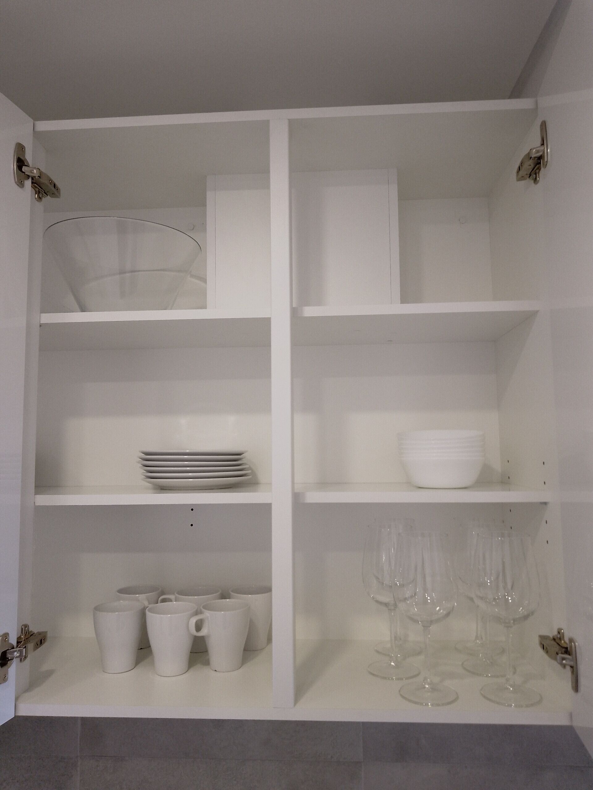 A white, open cabinet with shelves containing white plates, bowls, cups, and wine glasses.