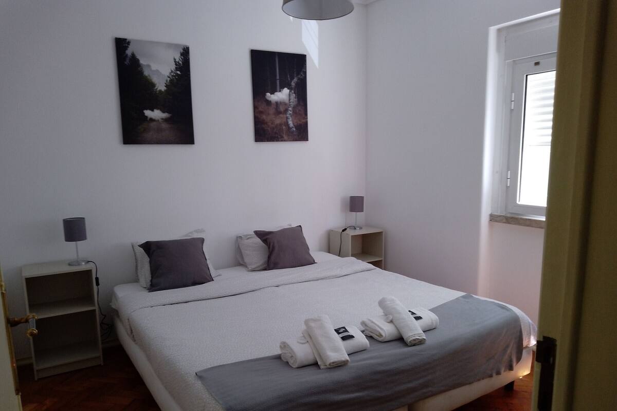 Una cama bien hecha con una manta gris, dos almohadas y toallas enrolladas. Hay dos mesitas de noche con lámparas, una ventana con persianas y dos cuadros en la pared.