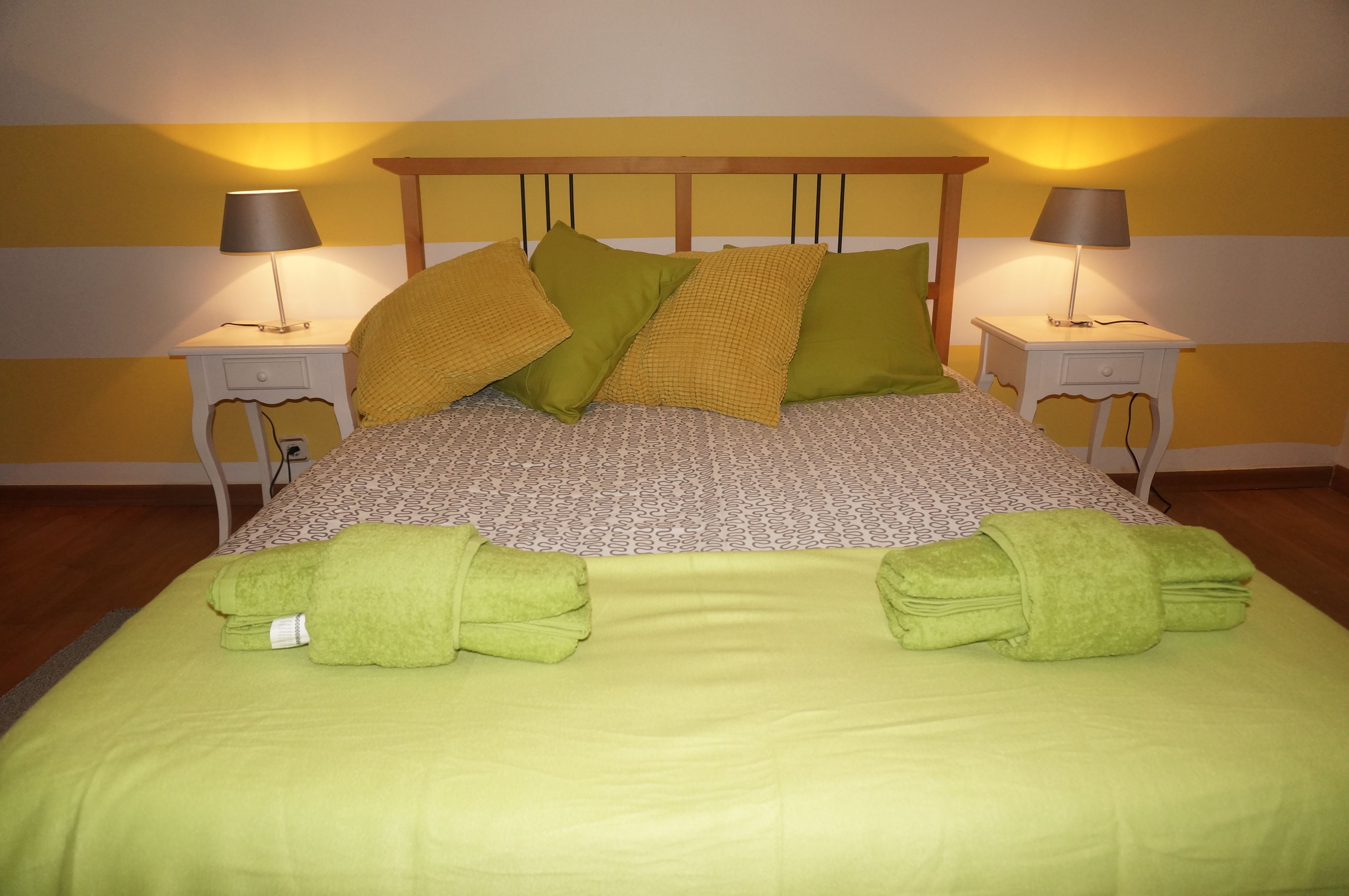 Una cama bien tendida con toallas verdes, una colcha estampada y dos lámparas de noche sobre mesitas blancas.