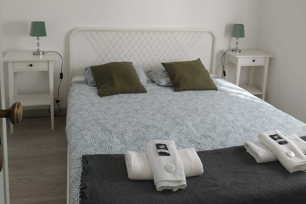 Una cama bien hecha con una colcha estampada y dos almohadas, flanqueada por dos mesitas de noche con lámparas, en una habitación sencilla con paredes blancas y suelo de madera.