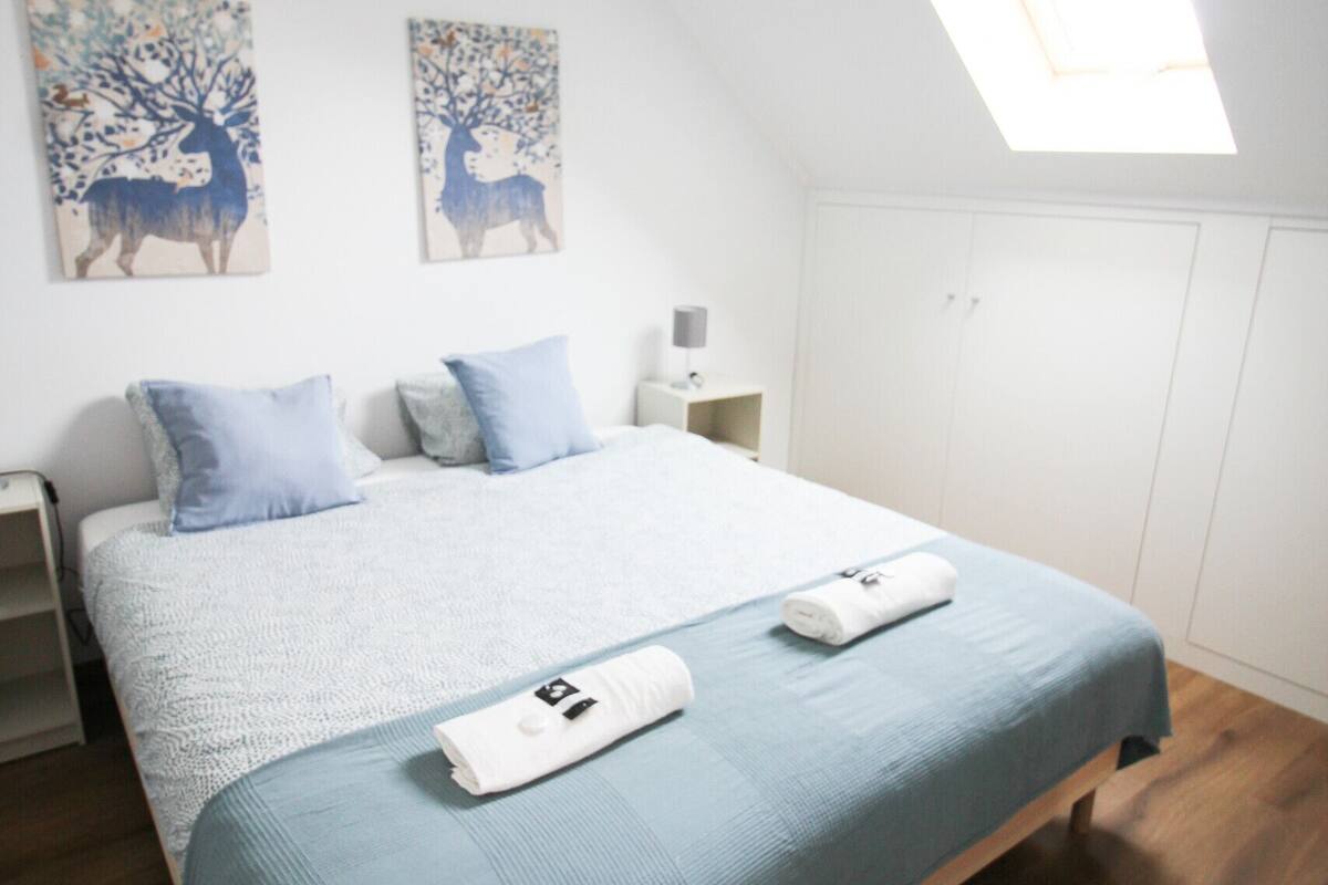 Un dormitorio con una cama grande, dos cuadros en la pared, un tragaluz y una mesita de noche con lámpara.