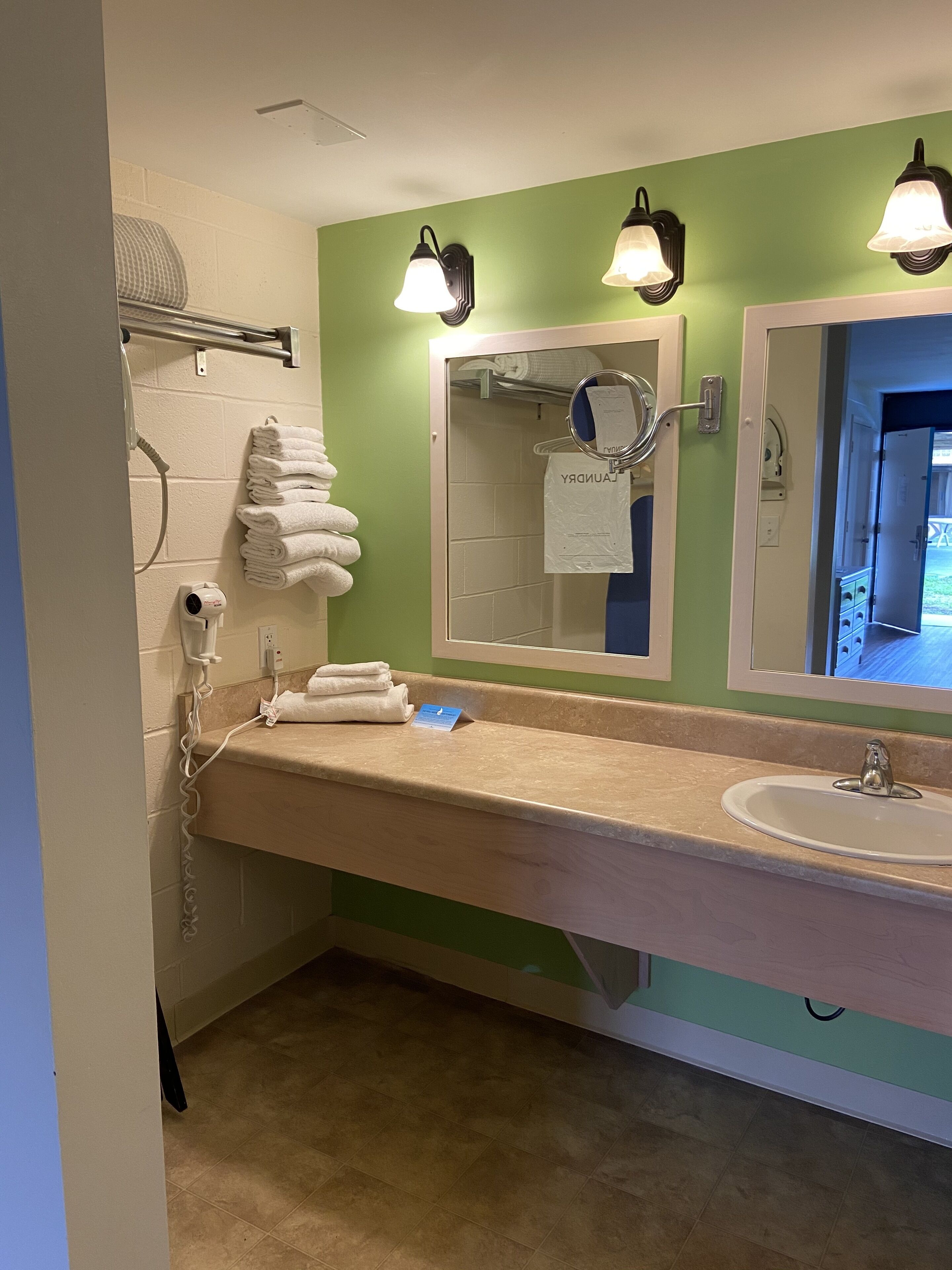 Un bagno con un piano di lavoro color beige, un lavandino bianco e una parete verde. Sono presenti due luci a parete, un portasciugamani con asciugamani piegati e un soffione doccia a mano.